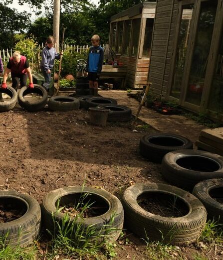 A teacher and two children stand in a wildlife garden, picking up tyres and digging in the soil. There is a big patch of soil in the middle, surrounded by black rubber tyres