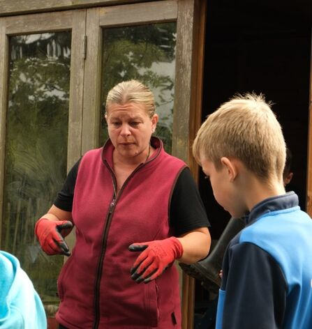 A teacher wearing a red fleecy gilet and red gardening gloves stands by a wooden door, speaking to a child wearing a blue jumper.