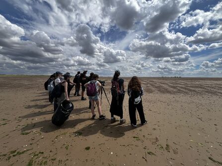 A group of young people, pictured from behind, stand on a beach with a camera on a tripod. It is a sunny day, with thick white clouds dotted around the sky.