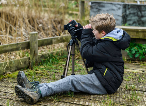 A blond-haired boy wearing a black coat and grey trousers sits on the ground looking through a camera on a tripod