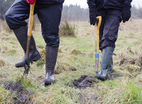 Two people seen from the waist down, digging with spades. They have waterproof clothing, gloves and wellie boots on. 