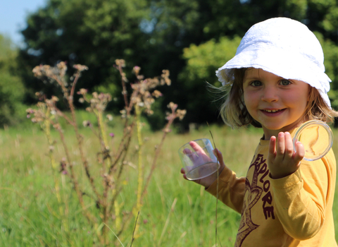 A child holds a bug pots and smiles in a meadow