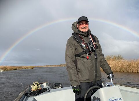 A smiling boat operator at Hickling standing in the boat wearing a thick coat and hat and with binoculars around his neck