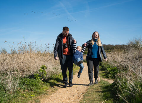 Two parents happily play with their young child as they walk down a path at sunny NWT Cley and Salthouse Marshes nature reserve