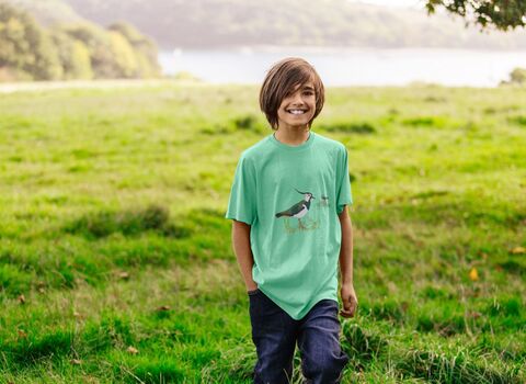 A young person walks through a field wearing a mint green loose-fitting t-shirt with a lapwing illustration printed on the front